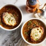 Elegant dining setup featuring a classic French Onion Soup in a traditional ceramic soup bowl with a toasted baguette slice and sprinkled parsley on top, accompanied by a silver spoon on a dark linen napkin.