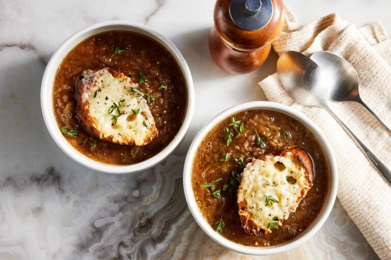 Elegant dining setup featuring a classic French Onion Soup in a traditional ceramic soup bowl with a toasted baguette slice and sprinkled parsley on top, accompanied by a silver spoon on a dark linen napkin.